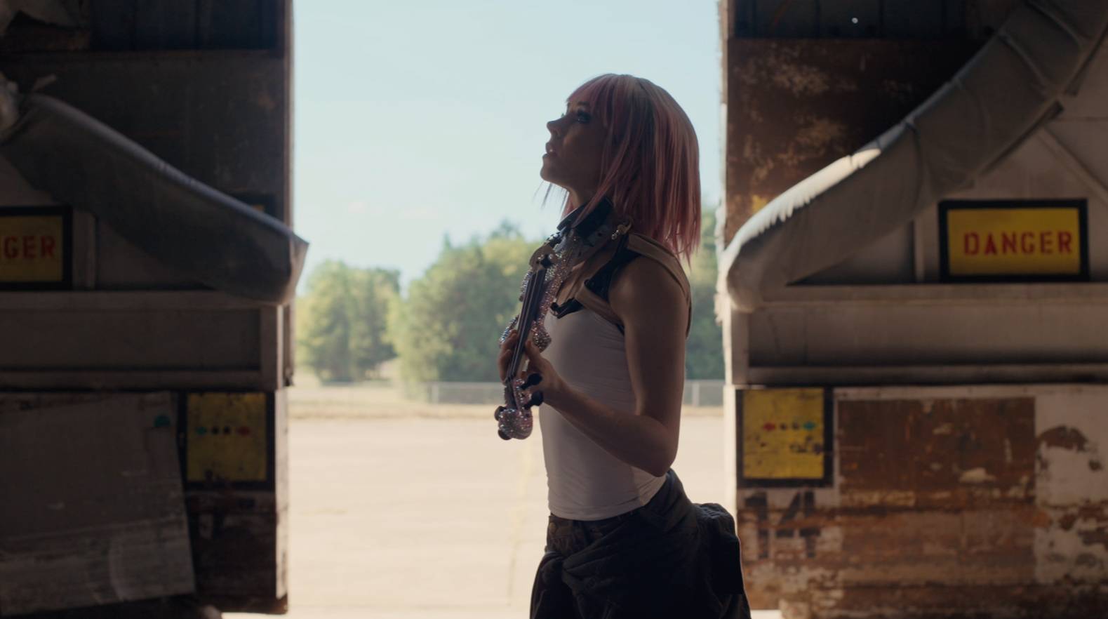 Lindsey Stirling in profile holding her jeweled violin between rusty industrial structures in ACE COMBAT 8 music video