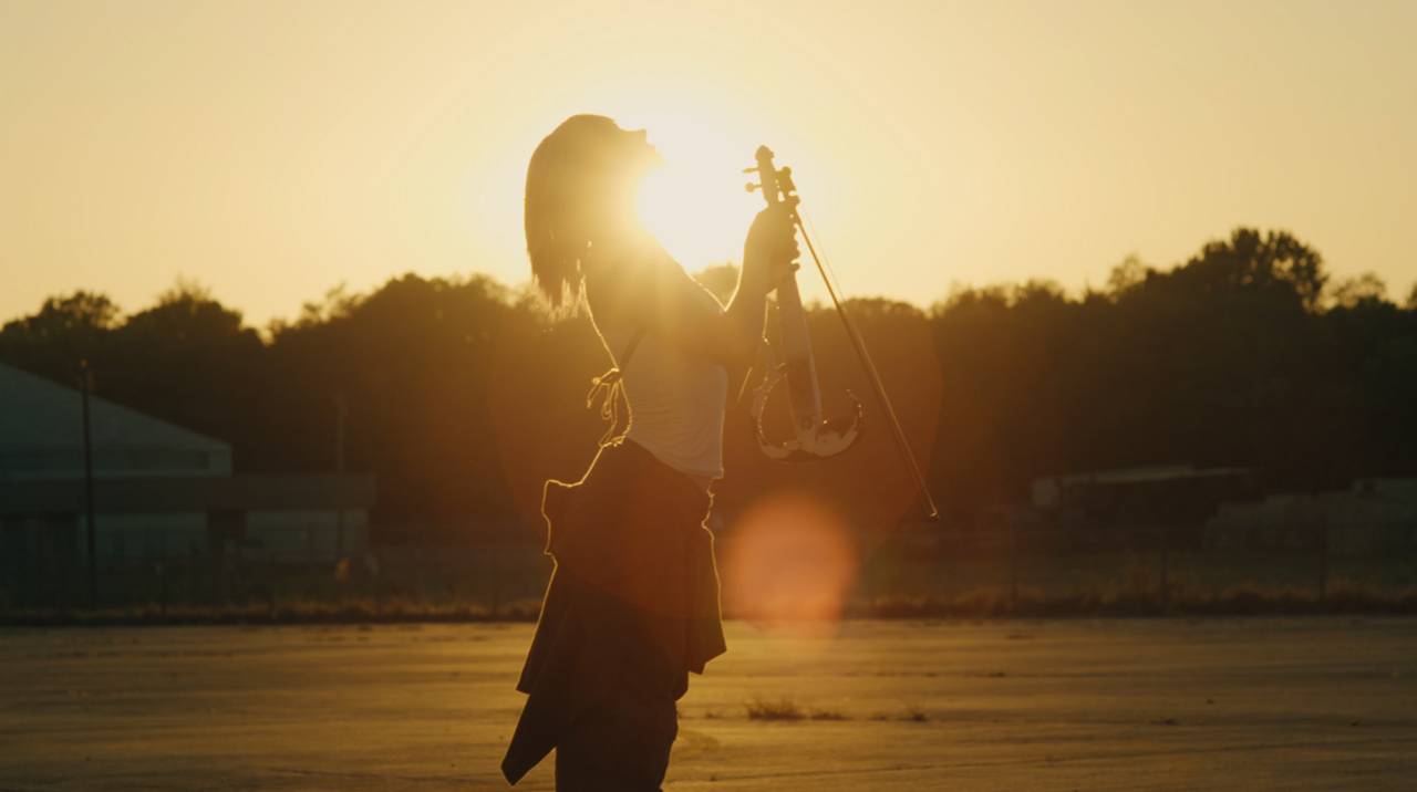 Silhouette of Lindsey Stirling raising her violin at sunset on an airfield in ACE COMBAT 8 music video